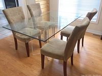 Full view of glass dining table with 4 suede chairs around it on hardwood floor near window. Shows transparent glass top, clear plexiglass legs, and chair upholstery color.
