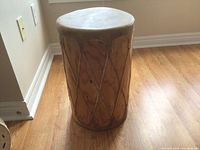 Front view of a large indigenous drum standing on hardwood floor, showing natural wooden frame and leather lace tensioning.