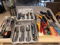 Wide view showing two trays with flatware sets and a tray with various kitchen tools and knives, including colored plastic handles and metal utensil holder with slotted spoons and spatulas.