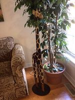Faux tree and wooden giraffe positioned beside a couch and window.