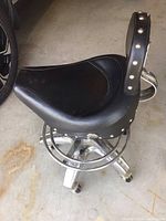 Side view showing black leather motorcycle-style seat with chrome base and footrest ring, metal studs along backrest edge.