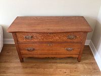 Front view of an old wooden dresser showing two drawers with ornate metal handles and keyholes, standing on a hardwood floor near a wall.