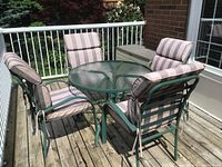 Photo of the full patio set on a wooden deck showing the round glass top table, four metal framed chairs with plastic webbing, each with a green and beige striped cushion attached.