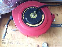 Photo showing red retractable air hose reel mounted on wooden surface with hose and brass fittings visible.