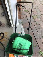 Green Scotts Turf Builder Edgeguard Mini manual spreader with metal handle and large treaded tires holding a green plastic poly tank sprayer with black hose and nozzle.