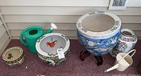 Shows all ceramic planters and two green watering cans from a distance, largest planter is blue and on a wooden stand, includes a planter with goldfish design and Asian vase with plate.