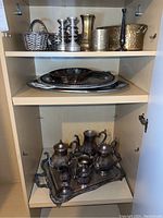 Full cabinet view showing silverplate coffee and tea server set with tray on bottom shelf, stacked platters in middle shelf, and mixed brass and silver items including baskets and candleholders on top shelf.