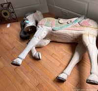 Photo showing full side view of carousel horse body lying on wooden floor with painted pastel green, pink and blue saddle detail, visible cracks in wood, and detached head with mane nearby.