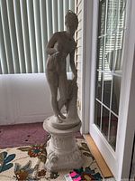 Full view of cement female figure sculpture standing on decorative base inside room with carpet and window blinds.