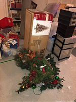 Boxed 7ft pre-lit flocked fir Christmas tree standing upright with various Christmas wreaths and garlands on the floor beside it in a basement setting.