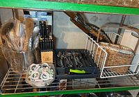 Overall view of metal shelf showing a stainless steel mixing bowl with wooden utensils, black-handled knives in wooden block, cutlery in plastic organizer tray, white metal basket with floral napkin rings, and wicker basket with floral liner on shelf below.