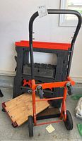 Metal dolly with wooden base, showing some rust and orange frame with wheels, next to black plastic sawhorses with red tops and a wooden creeper below.