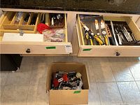 Wide view of two wooden drawers with multiple kitchen utensils, cook knives, and kitchen tools, plus a box with additional utensils on the floor.