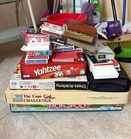 Stack of boxed board games Monopoly, The Cape Cod Challenge, Chess Academy, Yahtzee with multiple decks of cards and cases piled on top.