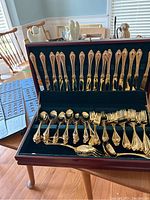 Opened flatware set case showing 16 place settings including knives and forks organized on slots with gold finish and ornate handles.