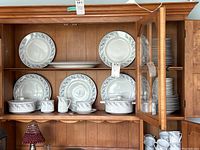Full set displayed in wooden china cabinet showing plates, bowls, cups, and serving pieces with blue floral border design.