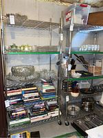 Two chrome metal shelving units with wire grid shelves, containing various glassware, dishes, books, and household items, located in basement.