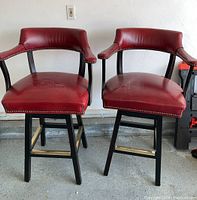 Two red vinyl upholstered Fairfield swivel bar chairs with nail head trim, brass footrests, and black wooden legs shown from front.