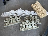 Five molded resin cherub and bird themed wall sculptures and shelves arranged on the floor, including cherub musical groups and decorative shelves.