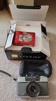 Nikon Coolpix L18 digital camera in red inside original box with manual, and Olympus-Pen EE-2 camera with black leather case in front.