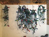 Six strings of outdoor Christmas lights with green wiring and multi-colored large bulb covers hanging against a wall.