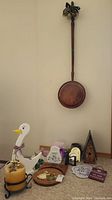 Full view of copper bed warmer hanging on wall with multiple smaller wood art pieces on floor beneath it including goose figure, candle, plaques, and birdhouse.