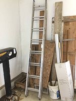 Full view of aluminum ladder leaning against wall surrounded by wooden boards and a bucket.