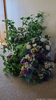 Cluster of faux plants and flowers displayed on carpeted floor against wall, showing large green leafy plants and flower arrangements in wicker and resin containers.