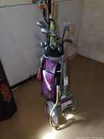 Full view of purple golf bag with multiple golf clubs including Spectra and MacGregor brands, push cart, and wooden tennis racket attached to cart handle.