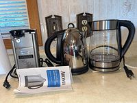 Photo showing Aroma electric kettle with manual, Kalorik glass electric kettle, and Cuisinart cast metal can opener on table.