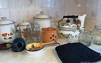Countertop kitchen items including 3 ceramic jars with floral designs, salt and pepper shakers, a black small ceramic pitcher, a ceramic spoon holder with floral design, a terracotta jar, a floral Asto kettle with brass handle, glass jar, and black oven mitt.