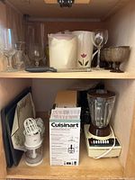 Photo showing Cuisinart Mini-Prep Plus food processor box, milk pitcher with pink flower design, assorted clear and dark glass stemware including goblets and wine glasses on wooden shelves.