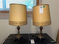 Pair of vintage table lamps on black surface in front of window showing fabric shades with floral trim and ornate metal bases.