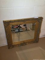 Full front view of ornate mirror leaning against white masonry wall showing intricate gold frame and bevelled glass