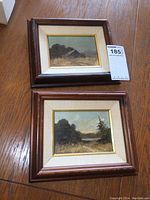 Pair of small framed oil paintings displayed on wooden table showing both paintings, frames, and price tag