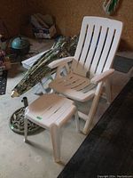 Full view of white resin folding chair with armrests and accompanying small resin side table, umbrella and wood table in background.