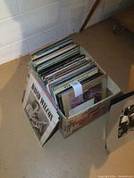 Cardboard box filled with approximately 80 vintage LP vinyl records stored vertically. Visible spine labels indicate diverse artists.