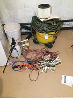 Photo of all items in the lot including the yellow 8 gallon shop vac, white garden sprayer, coiled green hose on top of the shop vac, and numerous extension cords bundled on floor