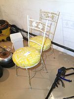 Pair of vintage metal bistro chairs with yellow vinyl round seats and floral metal backrests showing surface rust.