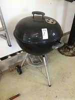 Full view of the black spherical Weber charcoal BBQ grill mounted on three metal legs, with two wheels and an ash catcher tray underneath.