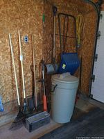 Photo showing yard tools including rakes, hoes, shovel, plastic trash can, extension cord, and blue snow shovel.
