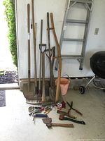 Photo showing multiple long-handled yard tools including shovels, hoes, pick axe, and scythe handle leaning against a wall, alongside smaller hand tools on the floor.