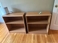 Two small wooden bookcases side by side on wooden floor near a wall and door, showing front view and shelves.