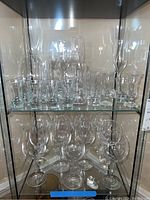 Two shelves of clear stemware and glassware inside a display cabinet, showing variety of types and sizes.