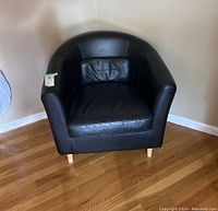 Front view of black leather-like club chair with curved back, attached lumbar pillow, and wooden legs on hardwood floor.