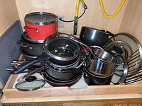 Photo showing a stacked collection of pots, fry pans, and lids including red ceramic casserole dish and black non-stick cookware