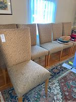 Six beige upholstered dining chairs with matching patterned fabric and wood legs, lined up in a room.