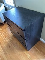 Overall left side angle view of the black three-drawer IKEA dresser on wooden floor near window.