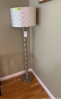 Full view of the floor lamp showing patterned lampshade, segmented brushed nickel stand, and round base on wood floor next to wall.