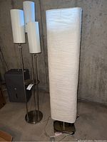 Two floor lamps against concrete wall. One with three white cylindrical shades and glass shelf, one with tall white rectangular paper-like shade.